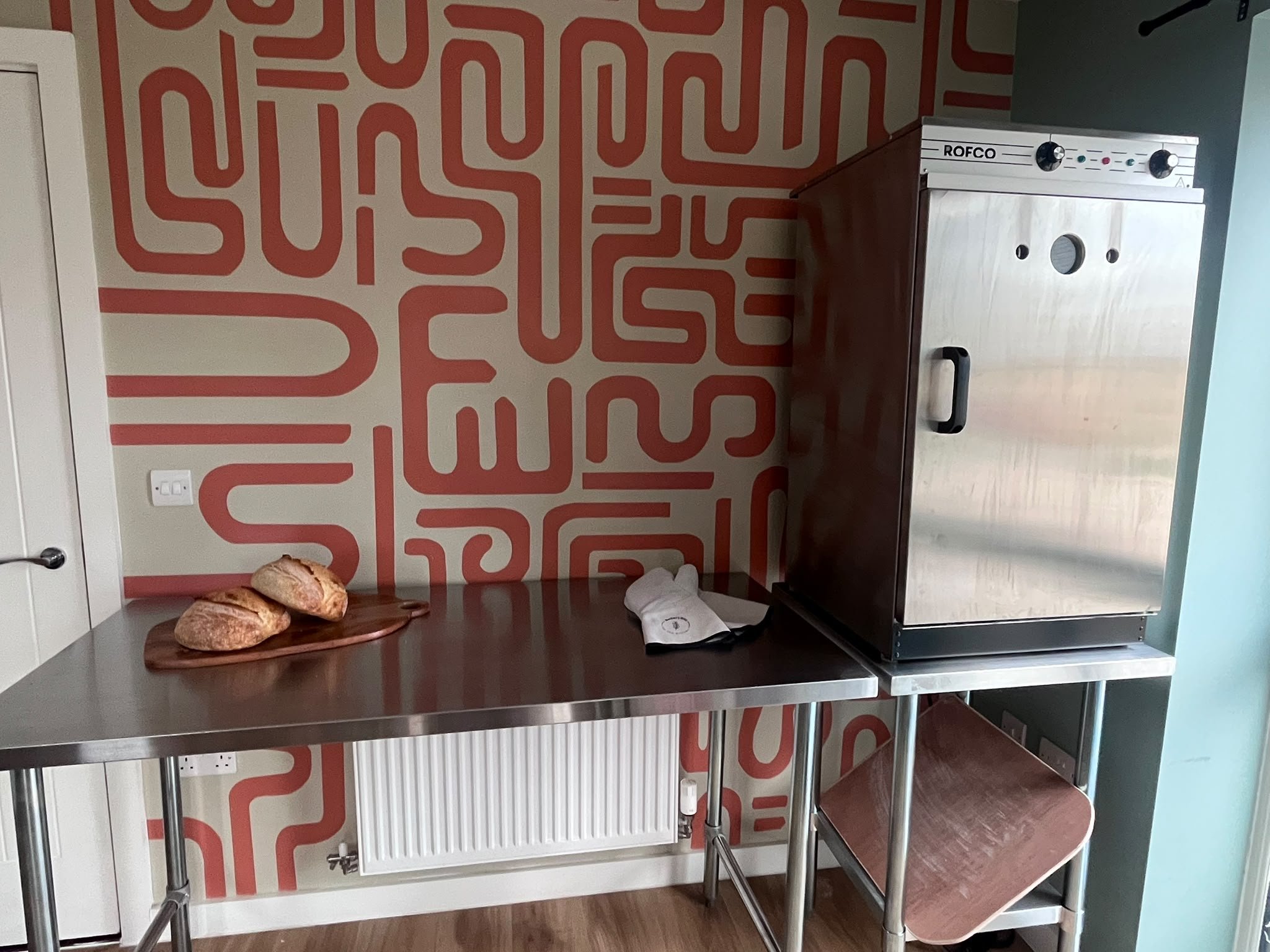 The bakery kitchen: two loaves on a wooden board on a stainless steel bench, oven gloves beside them, and the Rofco deck oven on its own stand, against a wall painted with a terracotta pattern.