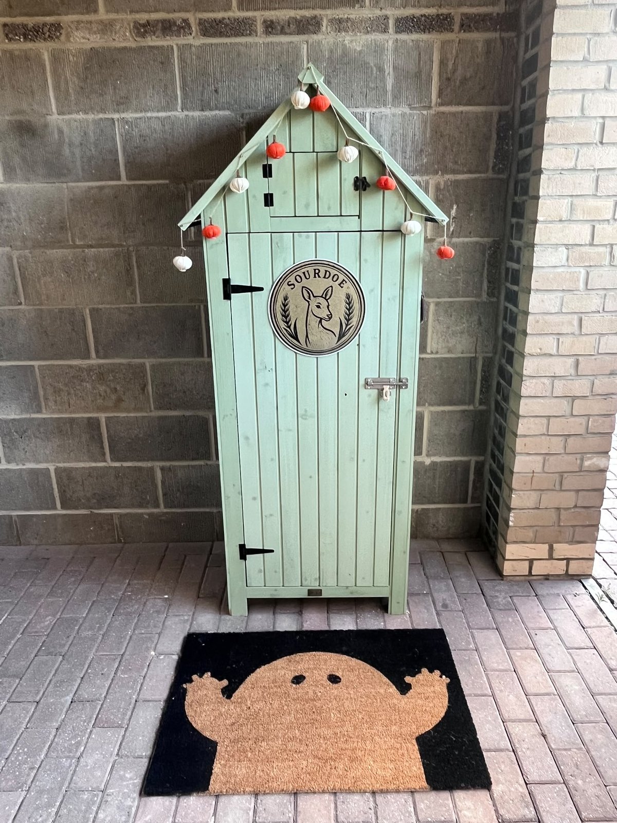 The bread shed: a narrow pale green wooden shed with a pitched roof and the round Sourdoe sign on its door, against a brick wall in the carport, with a doormat in front of it.